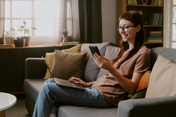 Obraz premium Caucasian young adult woman sitting on sofa using smartphone and laptop for online shopping smiling while browsing digital store in living room 