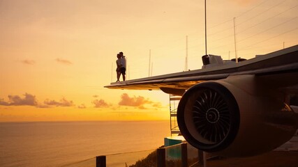 Romantic couple embraces on the wing of airplane at sunset, with the ocean sea view. Aircraft style hotel in Bali for vip luxury vacation and traveling.