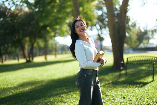 Asian woman student holding book smiling in park