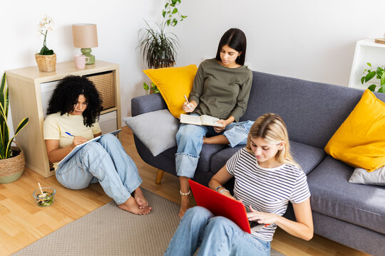 Three female students are collaborating on a project in their living room, using a laptop and notebooks, demonstrating teamwork and focused learning. Female friendship and youth concept