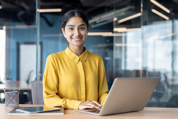 Portrait of a smiling young Indian businesswoman in a yellow shirt sitting at a desk in the office, working on a laptop and smiling at the camera
