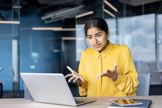 Young Indian businesswoman sitting at a desk in the office and talking casually to the laptop camera, gesturing with her hands
