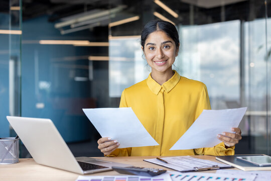 Portrait of a young successful Indian businesswoman sitting in the office at a desk with a laptop, holding documents in her hands and looking at the camera with a smile - Powered by Adobe
