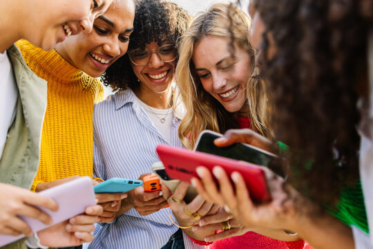 Diverse women standing together using smartphones and chatting happily outdoors. Young female friends connected through digital technology. Communication and friendship concept. - Powered by Adobe