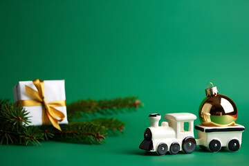 A minimalist Christmas scene with a small white gift, a toy train, and an ornament on a green background.