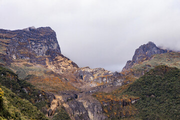 Picos escarpados del Nevado del Ruiz, Colombia: imponente valle andino con roca volcánica erosionada, vegetación de alta montaña verde y ocre bajo un cielo gris claro y nublado.