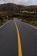 Carretera de montaña con línea amarilla sinuosa, atravesando el paisaje de páramo andino y acantilados rocosos bajo un cielo nublado, con un vehículo en la distancia.