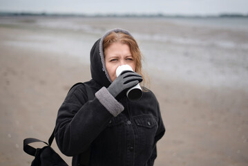 Woman resting after jogging on the beach and drinking hot tea with lemon. A woman in black coat and...