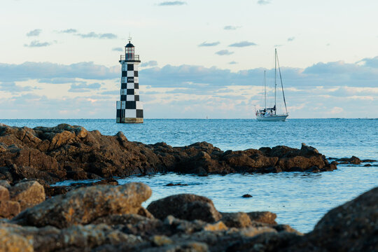 Checkered lighthouse and sailing boat beyond rocky shore, calm blue sea and soft evening light illustrating peaceful coastal navigation scene
