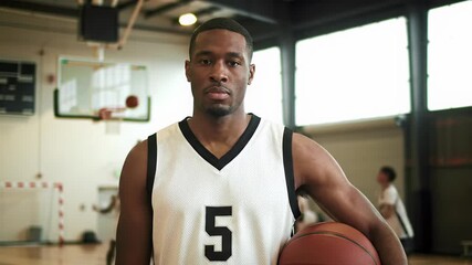 A young african american basketball player, mid-thirties, in a black and white jersey with '5', holding an orange basketball, stands poised on a court with blurred hoop. Concept of strength and - Powered by Adobe