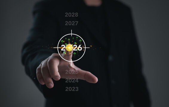 A hand points at the glowing 2026 target displayed on a digital interface, surrounded by upward green arrows and growth bars, symbolizing future goals, expansion, strategic planning, and rising perfor