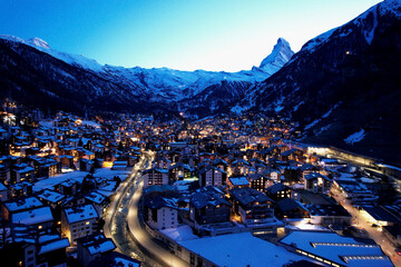 A stunning aerial drone view of Zermatt glowing at blue hour, with the iconic Matterhorn rising above snowy alpine peaks and winter lights of the village below.