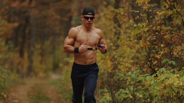 Young muscular shirtless man runs along path in an autumn forest with heart rate monitor on chest for control his load on body during his workout. Concept for effective training using technology