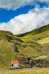 A rust-roofed refuge rests beside a cascading stream in Southern Iceland&rsquo;s mossy hills under a patchwork sky