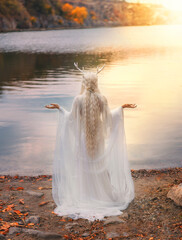 art photo real person, faerie fantasy woman is praying hands raised to sky by river lake water nature. sexy fairy tale elf girl long blonde hair white dress back, snow queen crown deer antlers on head