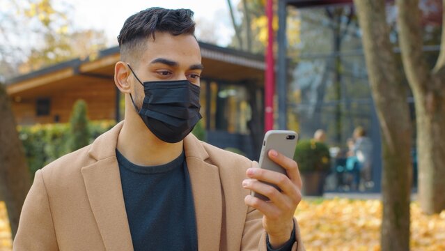 Hispanic man in face medical mask looking in mobile phone using app smartphone in park autumn fall season Arabian guy male chatting telephone social media chat outdoors positive online communication