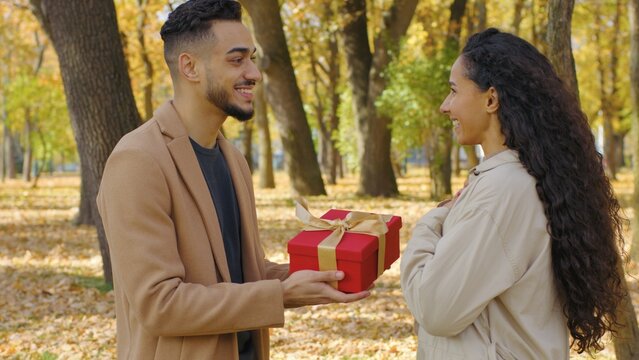 Hispanic happy couple lovely man giving birthday anniversary present gift box to woman in autumn city park hugging festive moment smiling girlfriend hug boyfriend surprise dating relationship love