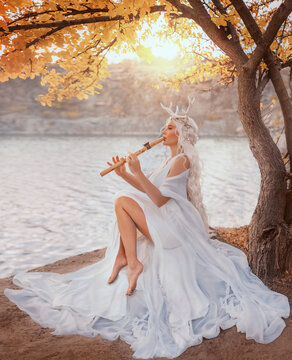 Portrait fantasy woman fairy faun plays a flute, sitting under yellow tree creating magical music. Myth happy sexy beauty face girl fae, long blonde hair white dress deer antlers on head. river rock 