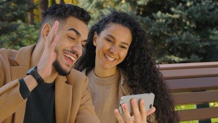 Happy hispanic couple talking video call with mobile phone in autumn city park sitting on bench family smiling Arabian woman man online talk conference with smartphone greeting vlog influencers chat