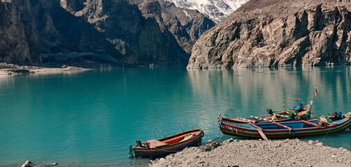 The vibrant Turquoise Waters of Attabad are captured, reflecting the colossal, rugged mountains that surround the lake. Two brightly painted wooden boats rest on the pebbled shore in the foreground,
