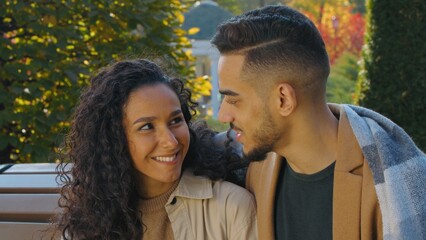 Cute Hispanic couple covered with plaid blanket date in autumn park on bench in city smiling...