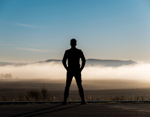 Silhouette of a Man Standing Outdoors, Overlooking a Misty Landscape at Sunrise with Mountains and Fog.
