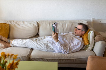 Obraz premium Relaxed middle-aged man in glasses reading a book while lying on a comfortable sofa