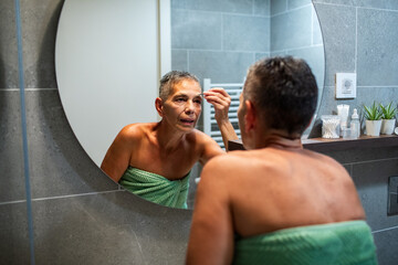 Mature woman grooming eyebrows in home bathroom mirror, focused
