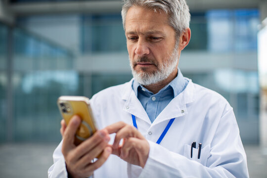 Mature doctor focused on smartphone outside hospital