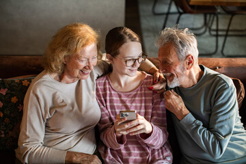 Senior grandparents and teen granddaughter laughing at smartphone on living room sofa