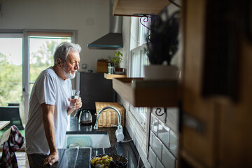 Senior man drinking water, thoughtful by window in home kitchen