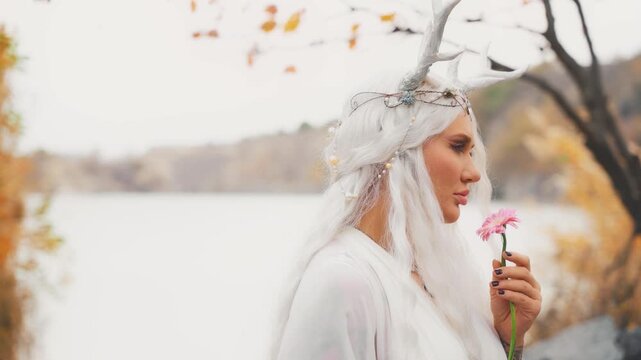 fantasy young elf woman holding pink gerbera flower. close-up beauty face sexy eyes lips fairy girl, deer antlers on head blonde ash hair white dress. myth faun pixie lady inhales floral scent aroma
