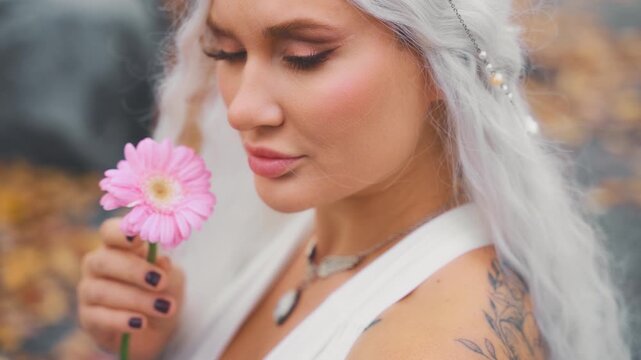 fantasy young elf woman holding pink gerbera flower. close-up beauty face sexy eyes lips fairy tale girl, blonde ash hair white dress trendy make-up. myth faun pixie sexy lady fashion model posing 4k