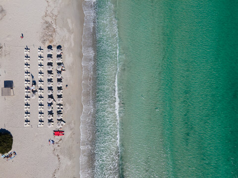 Aerial view of the shimmering turquoise waters meeting the pristine white sands of Cala Brandinchi beach, dotted with neat rows of sunbeds, San Teodoro, Sardinia, Italy.