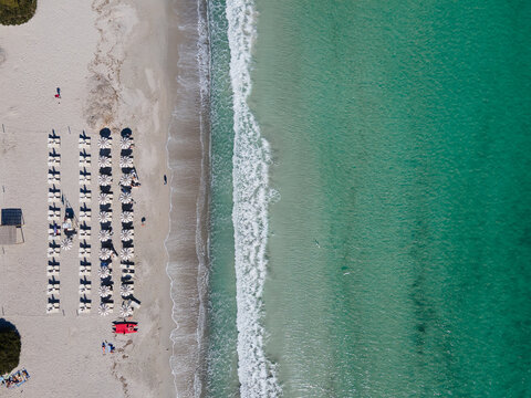 Aerial view of a pristine beach with rows of sunbeds meeting the turquoise sea with gentle waves, Cala Brandinchi, Sardinia, Italy.