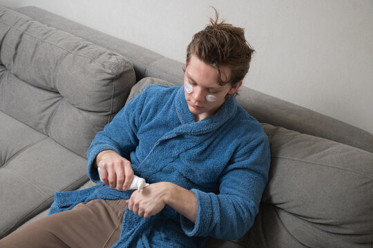 Young caucasian man in blue bathrobe applying skincare cream with under-eye patches while sitting on sofa during home selfcare routine. Mens Home Self-Care Routine