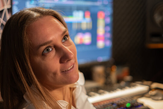 Female music producer with long hair, wearing a white shirt, engaged in creative work in a modern studio, surrounded by audio equipment and vibrant sound visuals