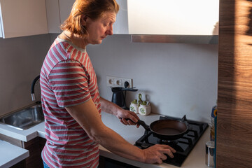 Caucasian man in a striped shirt cooking in a modern kitchen, using a frying pan on the stove, showcasing culinary skills and food preparation techniques