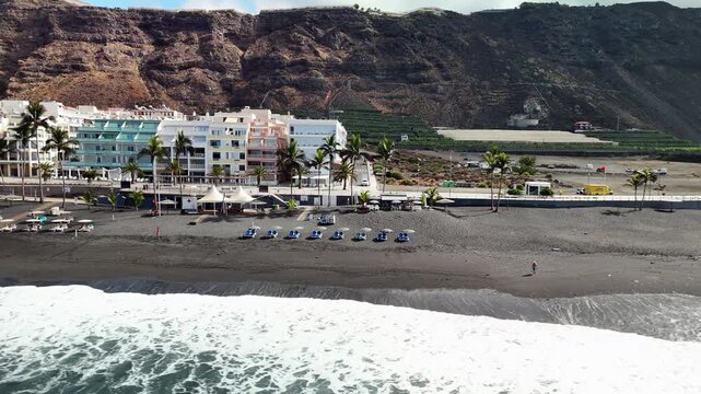 Low aerial drone pan over the Atlantic Ocean toward the black volcanic sand beach of Puerto Naos resort on La Palma, Spain.