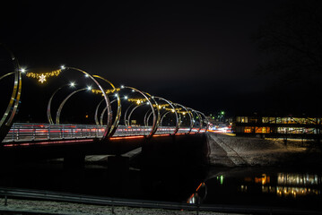 Night long exposure of a modern bridge with festive lights and traffic streaks over a dark river, winter banks and city glow.