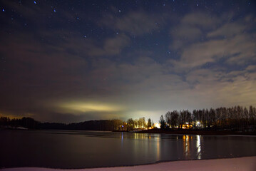 Starry winter night over a frozen lake, clouds drifting above silhouetted trees and distant lights reflecting on ice.