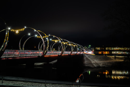 Night long exposure of a modern bridge with festive lights and traffic streaks over a dark river, winter banks and city glow.