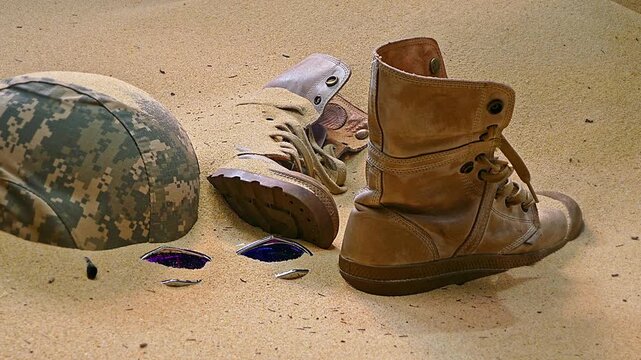 military helmet, a pair of worn combat boots, and a pair of broken sunglasses, partially buried in the sand. The arrangement suggests a narrative of someone having been in this harsh environment