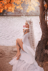 art photo real person fantasy young woman holding pink gerbera flower sittiing under tree. beauty face sexy eyes lips fairy girl, long blonde ash hair white elf dress trendy make-up. myth faun pixie