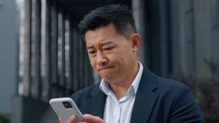 Middle-aged Asian businessman in city with phone sad Korean business man with mobile app failure upset worried looking in smartphone Chinese Japanese male entrepreneur outdoors using gadget problem