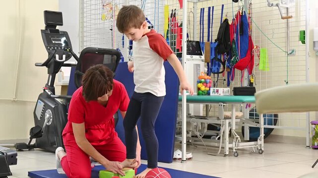 A boy doing exercises for flat feet, standing on his toes on an elevated platform, raising his toes under the supervision of a physiotherapist, undergoing rehabilitation