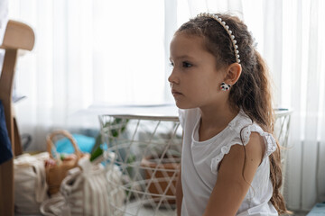 A young Hispanic girl sits cross-legged on a colorful mat at home. She has long brown hair and wears a white dress with pink ballet slippers. Natural light fills the room. © Alina