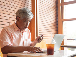 Concentrated senior Asian man holding a credit card while using a laptop for online shopping or banking at a cafe. Active elderly person showing digital literacy and independence in retirement.