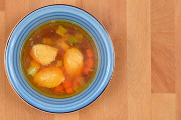 An overhead shot shows a blue rimmed bowl of clear broth soup with semolina dumplings and diced vegetables, resting on a light wooden background