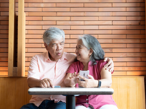 Active senior Asian couple using a smartphone together at a cafe or restaurant. The elderly man and woman are focused on the screen, showing digital literacy and connection in retirement.